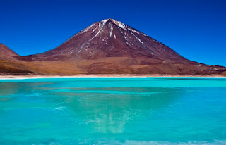 San Pedro de Atacama é uma pequena aldeia do Deserto do Atacama situada a 2.200 metros de altura ao longo das margens do Salar de Atacama, o maior lago de sal no Chile. O vulcão Licancabur ​com 5.920 metros está localizado a leste da cidade e domina a paisagem de San Pedro de Atacama e o Salar. Descrição: Foto do imponente vulcão em dia de céu azulão e limpo. Em primeiro plano, o lago de sal verde azulado que espelha o majestoso Licancabur com o cume nevado​. O cenário ao redor é ​erm​o ​com vegetação baixa e escassa.