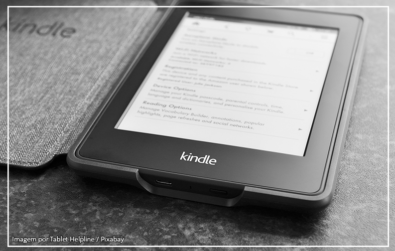 A imagem mostra um dispositivo Kindle sobre uma mesa. O dispositivo é preto, retangular e está com a tela ligada, mostrando um texto. Abaixo é possível ver a palavra Kindle escrita nele