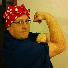 The author, in his Halloween costume as Rosie the Riveter, wearing the dark blue shirt and red bandana with white dots, striking the traditional 'We Can Do It!' pose with one arm flexed as the other arm rolls up the shirt sleeve.