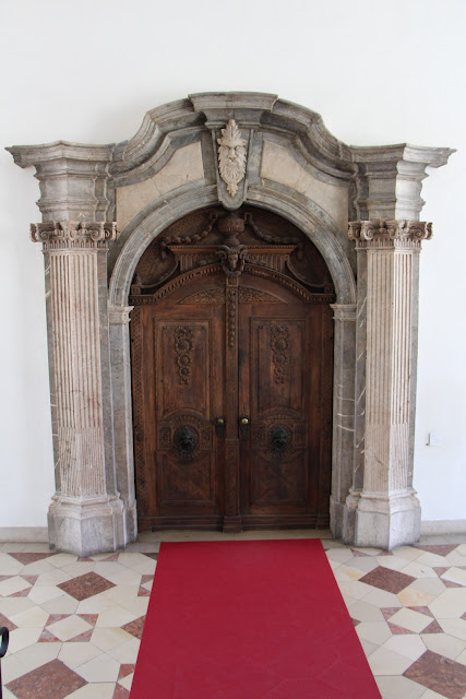 Photo of a door. The door itself is medieval or Renaissance, German, and made of wood. It's elaborately carved, with rosettes carved vertically, a goat's head, vegetation, and more. It is set within a carved stone surround that includes a green man (maybe) and fluted columns on either side. It's set in a wall and the floor in front of it is elaborately geometric, in white and a light red.