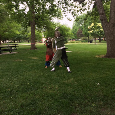 Two people, one much shorter than the other, in medieval clothing and armour. They're swinging rattan swords at each other in a green park with tall trees; the taller person in green and white has a shield, and the shorter person in red and blue has only a sword.