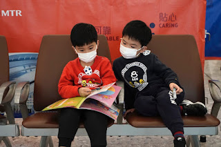 A boy in red t-shirt wearing a mask reading a book sitting close to another boy wearing a mask spreading coronavirus through human contact