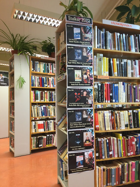 vaade raamatukogu ilukirjanduse osakonna keskelt. Ülepea kerkivad raamaturiiulid on raamatuid tihedalt täis, riiulite kohal haljendavad toataimed. Ühe riiuli külg on kaetud plakatitega, millel on tekst ja portreed 2023 aasta ulmeauhinna stalker võitjatest.