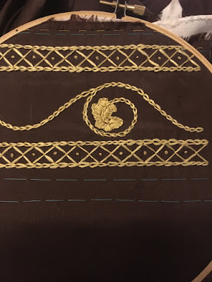 A piece of dark brown silk stretched in a wooden embroidery hoop, with two narrow bands of diamond embroidery flanking a chain-stitched sine curve with a single curl and oak leaf springing from it.