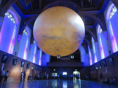 Photo of large red-brown sphere hanging in a large, hall, with tall windows covered by blinds on each side.