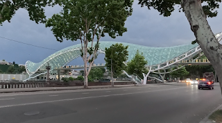 A glowing arc of glass and steel spanning the Kura River, illuminated softly against the cloudy skyline of Tbilisi