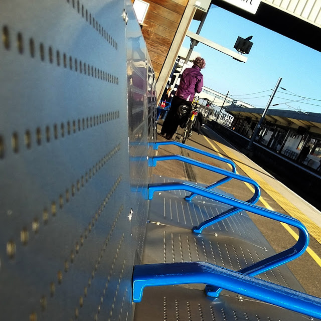 Looking along an unpainted steel railway 'bench', with blue seat dividers.  The arm of the bench is angled, and so the camera resting on it has captured a tilted, rather sideways, view along the platform and across the station.