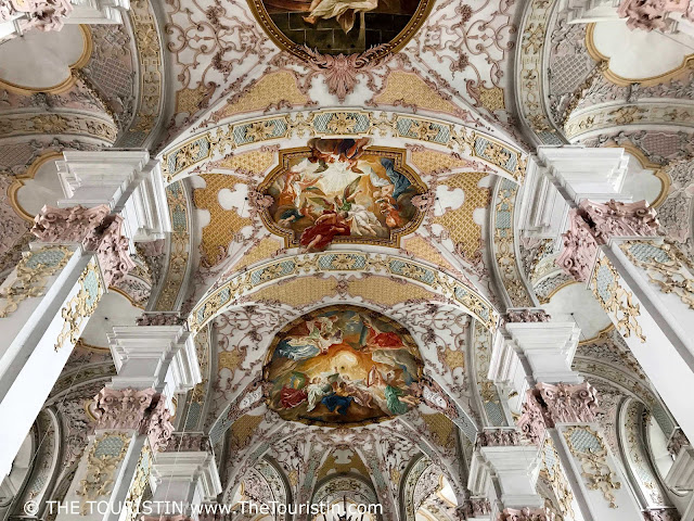 An elaborately decorated ceiling painting with a groups of people dressed differently in coloured robes and tunics, painted in pastel colours ranging from pink, light blue, and yellow, to red to gold.