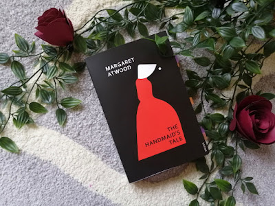 A book is on top of some leaves and two flowers. The book cover has a black background with a silhouette of woman where her white bonnet and red dress are emphasised. The text reads 'Margaret Atwood' 'The Handmaid's Tale'