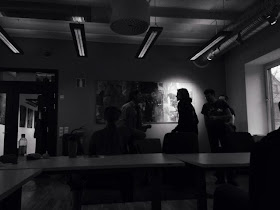 [a photo of a classroom in black and white. The tables are empty but some people have gathered at the back of the room. You can mostly see their dark shadows.]