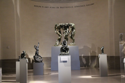 Photograph. Hall at the Palace of the Legion of Honor with multiple bronze statues by Rodin, all on white pedestals, including a grouping of three figures at the rear of the photo, in front of a wall.