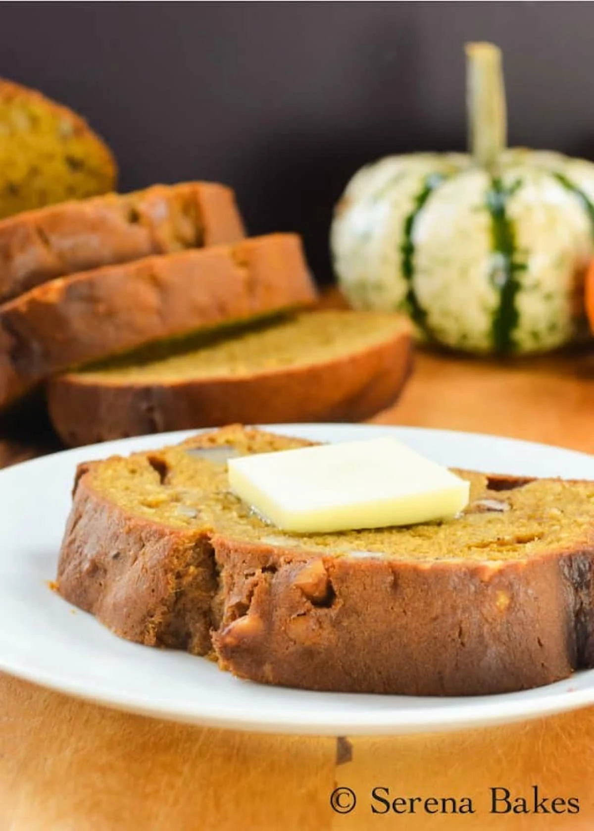 A slice of Pumpkin Bread topped with a pat of butter on a white plate with a sliced loaf of Pumpkin Bread in the background.