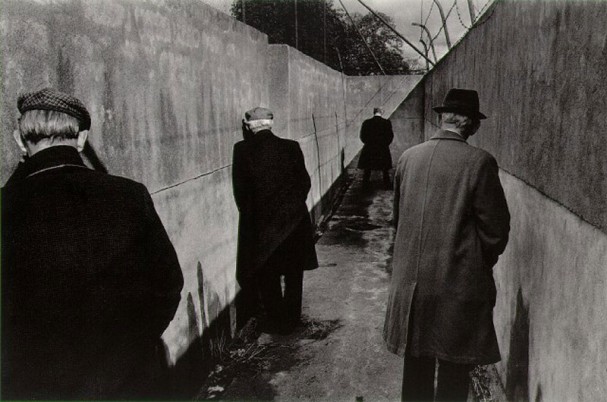 Fotografia em preto e branco de um beco sem saída. Há quatro homens de costas. Eles vestem sobretudos e chapéis e seus cabelos parecem ser brancos.