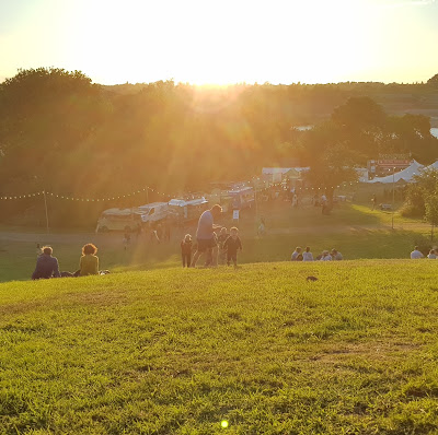 Autumnal evening view from the top of a hill, as the sun sets over a festival scene, with people sitting on the hill below