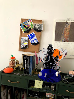 an image of the top half of the desk with decorations for halloween including a beistle paper stand up haunted house centerpiece, a pipe cleaner pumpkin and bat clips on the bulletin board above