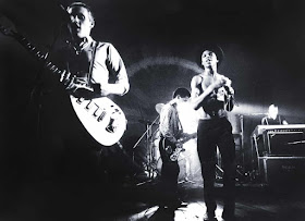 In the foreground, Dave Wakeling plays guitar and sings into a microphone, while a shirtless Ranking Roger holds a mic and dances next to him.