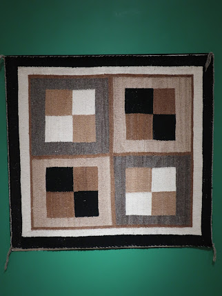 Photo of a knitted or crocheted artwork. Squares in various shades are knitted or sewn together to make patterns in shades of brown, tan, beige, black, and white. The work is mounted on a green background.