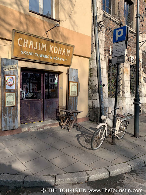 Cream coloured bicycle leaning against the post of a street sign. Opposite a wooden coffee house table in front of the entrance of a restaurant with the name over its glass door.