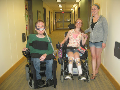 Me seated in a powerchair between my friend David and his sister Megan who is also my friend. David is also seated in a wheelchair, wearing a ventilator mask under his nose. Megan is standing beside me. All are smiling in the dorm corridor