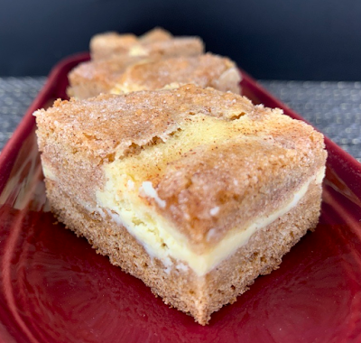 Snickerdoodle Cheesecake Bars: a base layer of snickerdoodle dough topped with a cheesecake filling and a topping of additional cheesecake dough. Photographed on a burgundy oblong plate.