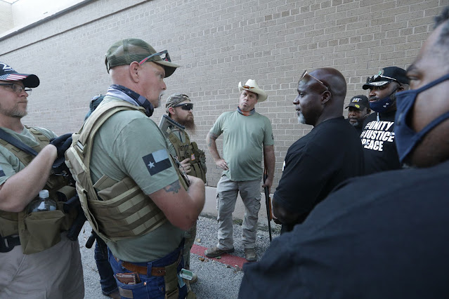 Five white men in matching t-shirts, at least three of whom also wear military-style tactical vests and appear to be armed, stand together and exchange looks with four black men who stand across from them, wearing matching T-shirts of a different design bearing the words “#UNITY #JUSTICE #PEACE.” What are they thinking about this encounter?