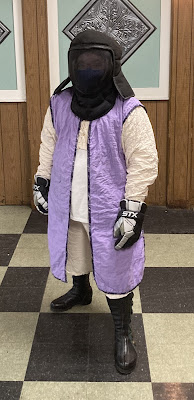 A person is standing with their hands held loosely at their sides. They are wearing a black fencing mask/hood, black gloves, black boots, and a white/lavender ensemble of a jacket, tunic, vest, and shorts.