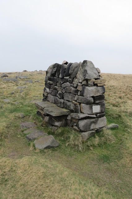 A memorial bench on the moor, built like a dry stone wall with two flat stones halfway up laid to form a seat.