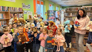 Zdjęcie grupowe na którym znajdują się dzieci z  przedszkola wraz z paniami bibliotekarkami. Wszyscy stoją razem przed ladą biblioteczną i regałami z książkami. W tle widać też pleksę na której przyklejone są kolorowe naklejki przedstawiające jesienne postacie np. wiewiórki i lisa i listki. Dzieci trzymają w dłoniach pluszowe misie.