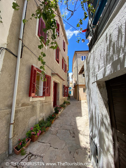 Three-storeyed houses decorated with red shutters and flower pots on a narrow lane that runs towards a three-storeyed house with blue shutters and a blue front door.