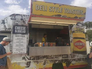 Open food stand with black and white old fashioned photos. On the left it says "Cheese & Bread" with the menu underneath. In the center is the open window to order food. A person in yellow is standing behind the counter. There are 6 different soda bottles in front of the counter. To the right of the open window is an image of a container of Vegemite that covers the side of the struck. Above the window is a yellow sign that says in white letters "Deli Style Toasties"
