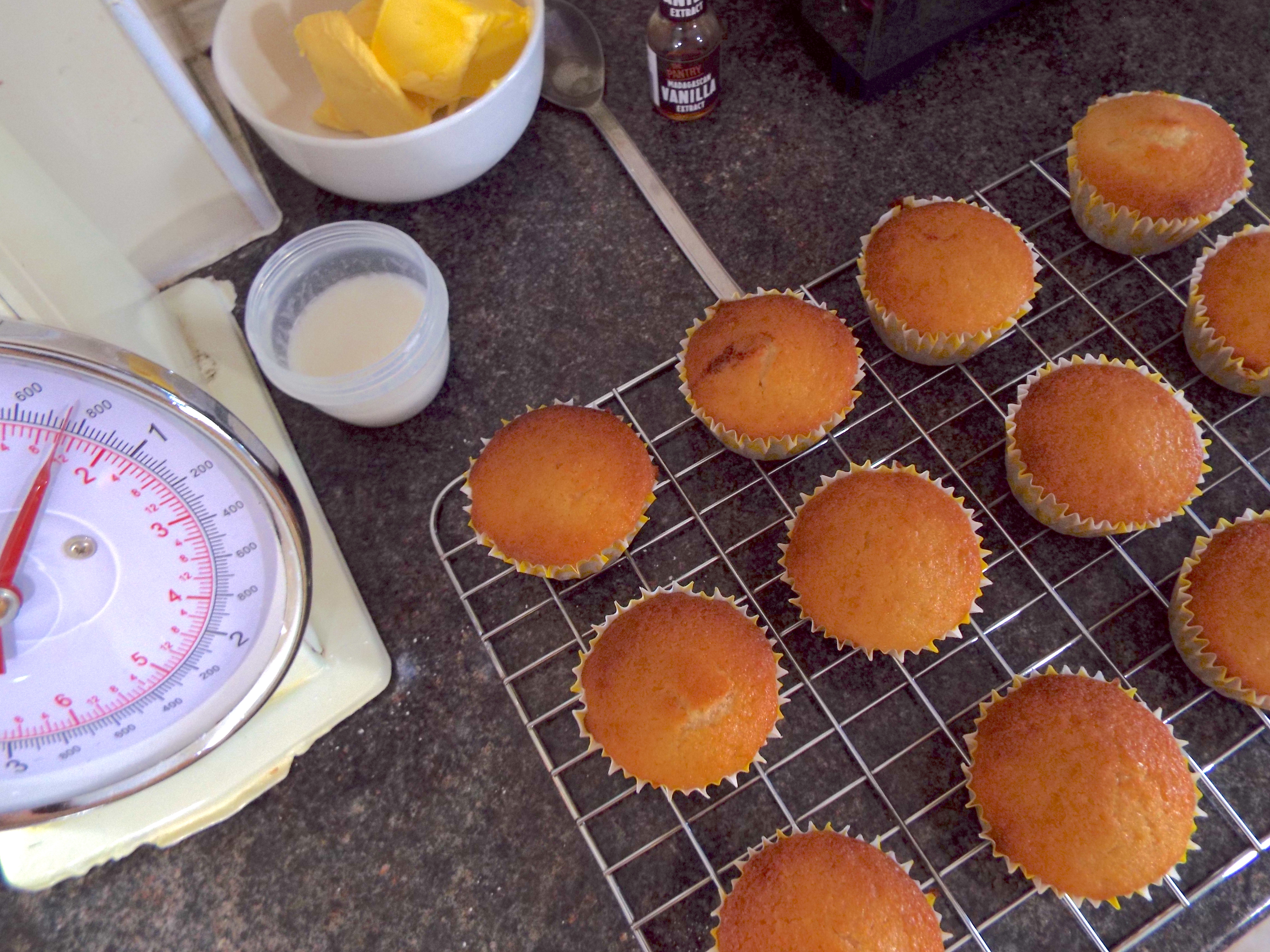 Freshly baked cupcakes on kitchen counter, beside cream old fashioned scales, butter and icing sugar.