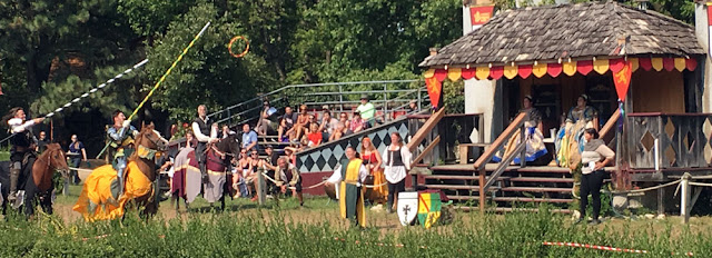 A panorama of two mounted men holding barber-poled lances, one in gold and green and the other in black and white, aiming to catch a small gold-and-red hoop tossed in the air in front of a plaster-and-timber viewing platform on which the queen and her ladies, in Tudor garb, sit watching. Various attendants in Renaissance clothing stand at the sides of the list field, and a small set of stands to the left of the viewing platform are full of people in modern clothing.