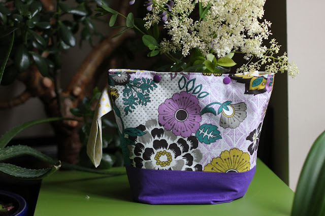 Purple floral project bag with purple boxed bottom and yellow triangle side loop and purple snap buttons on top. On an apple green table in front of white Japanese lilac and beloved house plants.