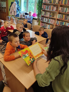 Pani bibliotekarka siedzi naprzeciwko grupy przedszkolaków i czyta im książkę o tym jak fenek sadzi drzewko w sadzie. Dzieci siedzą przy bezowych drewnianych stolikach wokół regały z ksiązkami.