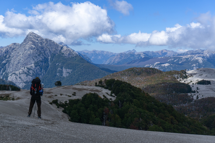 A man is walking on a trail on the mountains. He is carrying a backpack and looking down in the landscape.