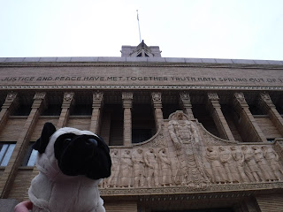 a plush pug appears in front of an elaborately carved building front featuring many pillars, many carved men, and the words "justice and peace have met together. truth hath sprung out of" visible