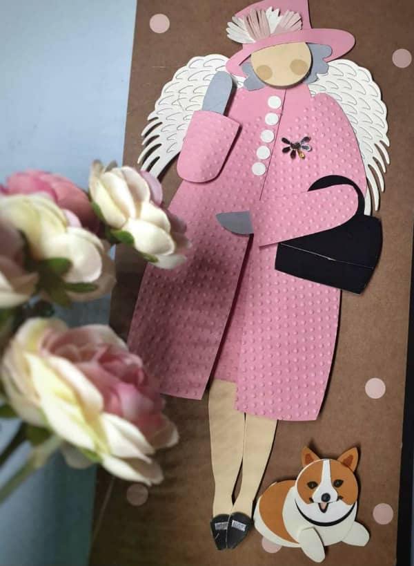 Paper cut figure of Queen Eizbeth with wings and a corgi next to a bouquet of roses