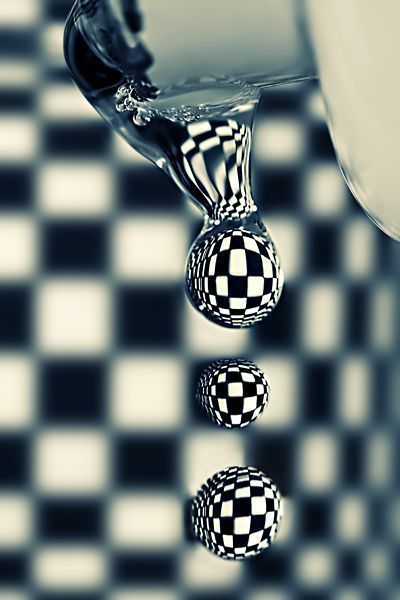 Close up of drops of water falling from a tap in a room with black and white checked walls. The black and white check is reflected perfectly in the droplets of water. Photo take by Sortvind on Deviant Art