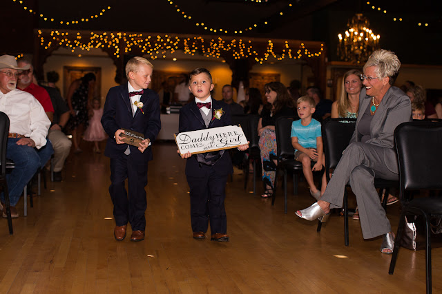  Cassie and Wyatt tied the knot at The Palace Event Center, in Yukon, OK, on July 15, 2017.   The venue’s magnificent wood entry doorways, outside rock façade, and adjacent country landscape provided the perfect backdrop to photograph the first look and pictures of the jovial wedding party.  The venue was spacious, which allowed us to sneak in a few pre-dressed shots of the girls in their “Wedding Tribe” t-shirts.     Cassie’s elegant, form-fitting, strapless, satin wedding dress overlaid with lace and sequins looked like it was made especially for her.  Wyatt was quite the Knight in Shining Armor dressed in his navy suit, grey vest, and burgundy tie.  The four beautiful bridesmaids wore matching full-length, flowing, burgundy gowns, slit at the knee.  Cassie’s gorgeous bouquet was composed of baby’s breath, peonies and ranunculus in rich hues of vibrant burgundy, red, pink, burnt orange, and yellow gold.  To add to the excitement of the day, their son had a special assignment—carrying the sign down the aisle that stated, “Daddy, Here Comes Our Girl.”     The venue’s grand ballroom, with it’s beautiful chandeliers and romantic lighting, was the perfect backdrop for the couple’s reception.  Candlelight, flowers, and twinkle lights adorned the nuptial stage, and after the vow exchange, Cassie and Wyatt participated in their first dance.  Guests were invited to enjoy the buffet, the cutting of the cake, and the toasts by the best man and bridesmaid.  Chairs were quickly removed from the dance floor, and while almost everyone spent the rest of the afternoon relaxing, dancing and socializing, there was a particularly fun-loving group of guests who decorated the bride and groom’s Jeep with sayings, crepe paper, and a car filled-to-the-brim with balloons!        Cassie stated that the best part of the wedding was being able to see so many family and friends at the reception having fun and enjoying themselves.  She said, “If we had it to do over, we wouldn’t do anything differently…it was perfect for both of us!”     It was a pleasure to capture the special moments of this memorable wedding day for Cassie and Wyatt, and we wish them a lifetime of love and happiness. 