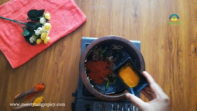 Red chili powder and turmeric powder being sautéed with onions, green chilies, and ginger in an earthen pot.