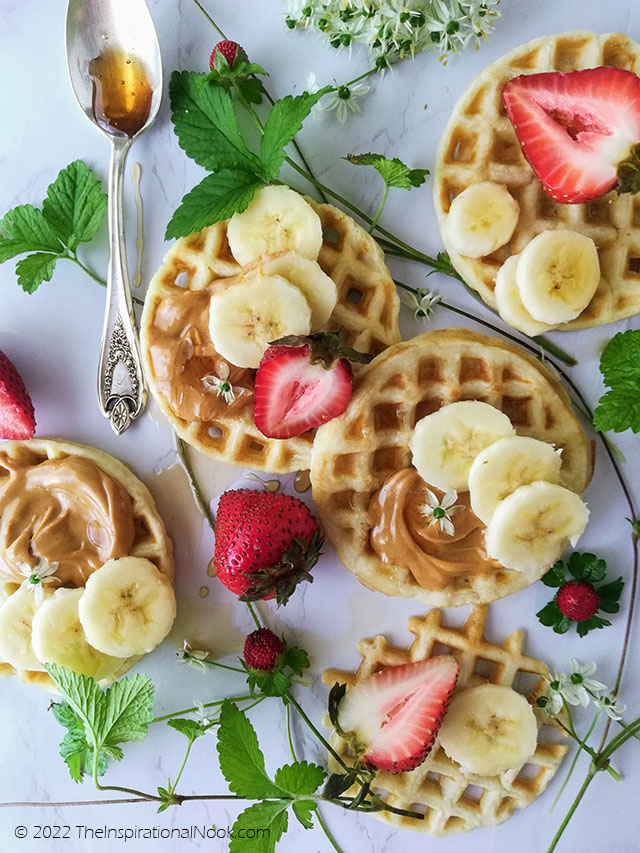 Round mini waffles with peanut butter, strawberries, bananas and a spoonful of honey, surrounded by strawberry leaves and wild strawberries.