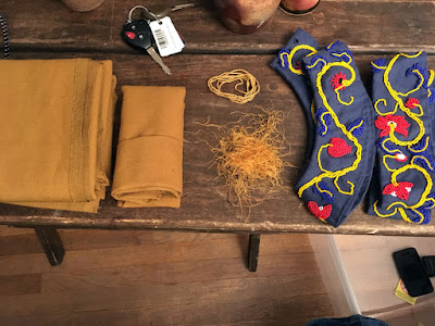 A photo from above of two folded stacks of mustard-brown fabric, a pile of mustardy thread scraps and a coil of bright-yellow cord, a curved blue collar with yellow, red, and blue beading in an undulating vine, and a straight strip of blue fabric with yellow, red, and blue beading in an undulating vine, all on a worn dark wood table.