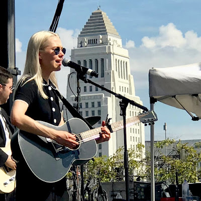 Woman with blond hair in black dress playing guitar and singing with LA City Hall in the background