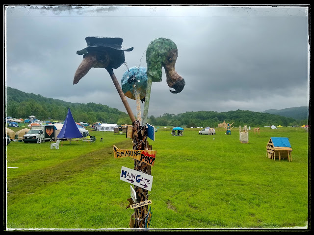 A tall signpost capped by three oversized bird heads, in a grassy field