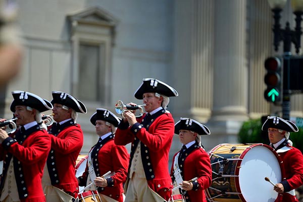 men in red coats and tricornered hats march in formation playing drums and bugles