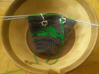 Double-knitting in the round on dpns.  The yarns are green and dark brown.  The increases for the thumb gusset are marked with silver dolphin stitch markers, and the work is sitting in a wooden yarn bowl.