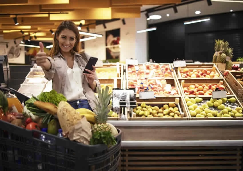 Imagem mostra uma mulher em um supermercado sorrindo com um smartphone em uma das mãos com a outra dando um like perto de um carrinho lotado de supermercado.