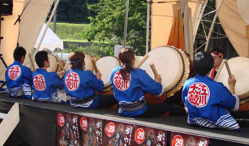 Members of a traditional taiko drumming group wearing happi coats sporting the group's name on the back. In this shot the three male drummers wear their happi loose, and the two females use an obi.