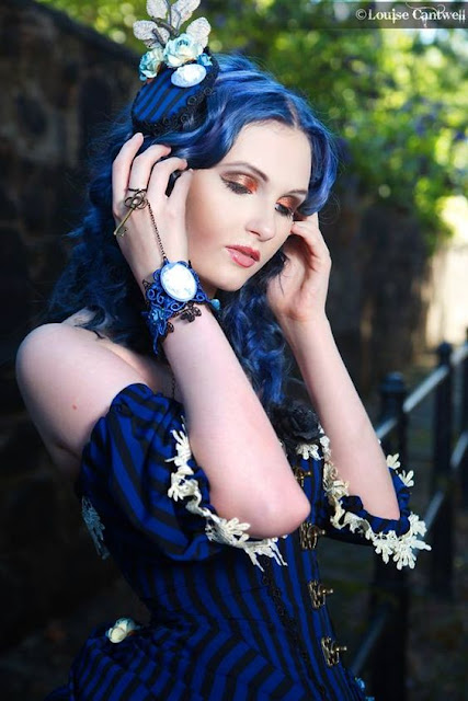 Steampunk woman in black and blue striped corset/dress. She has blue hair and bronze/copper eyshadow, blue gothic jewelry, and a blue fascinator. Subtle beautiful makeup.