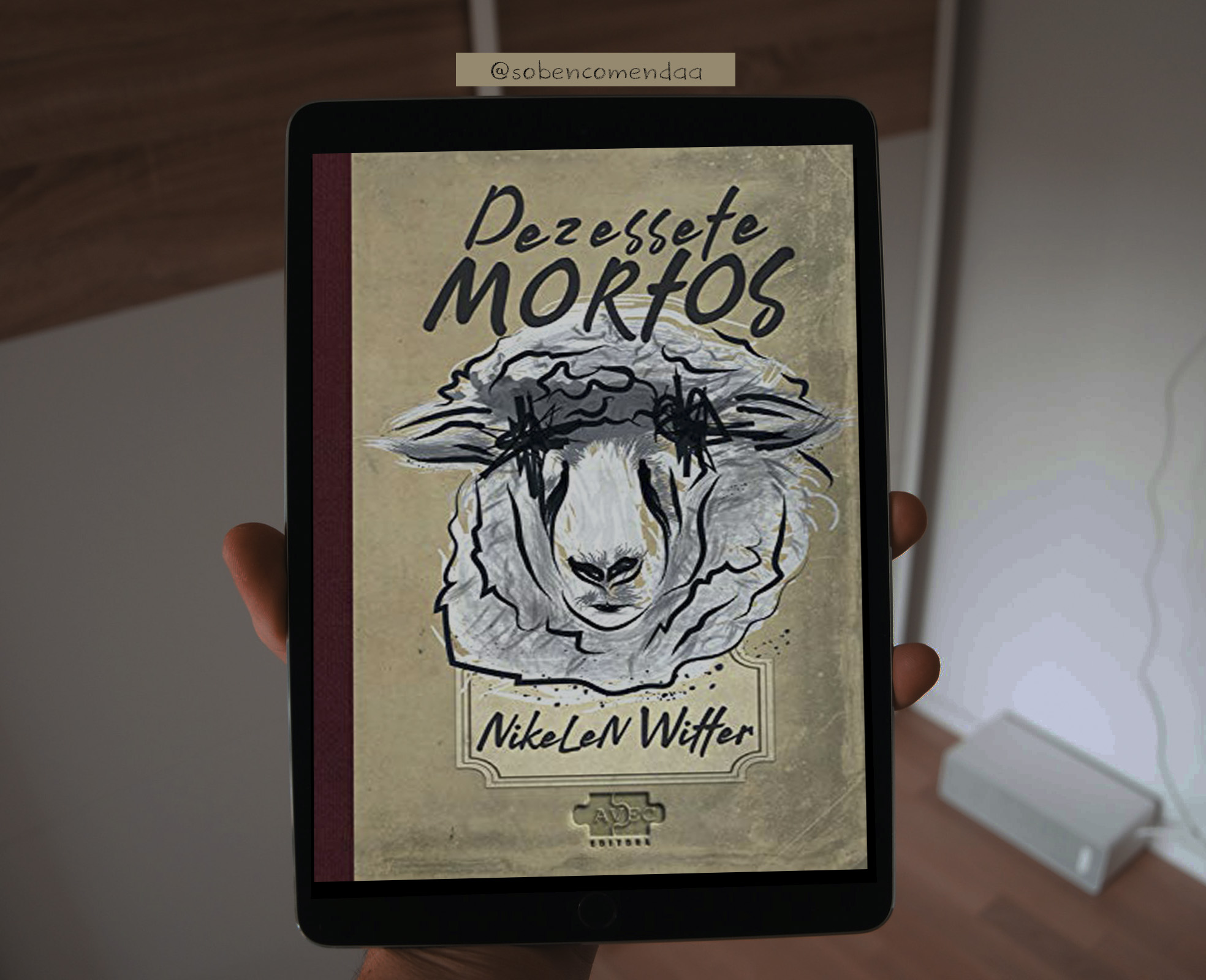 Imagem de uma mão segurando um tablet, com a imagem da capa do livro Dezessete Mortos, dentro de uma sala. Ao fundo uma parede branca, com o chão aparecendo, de madeira.